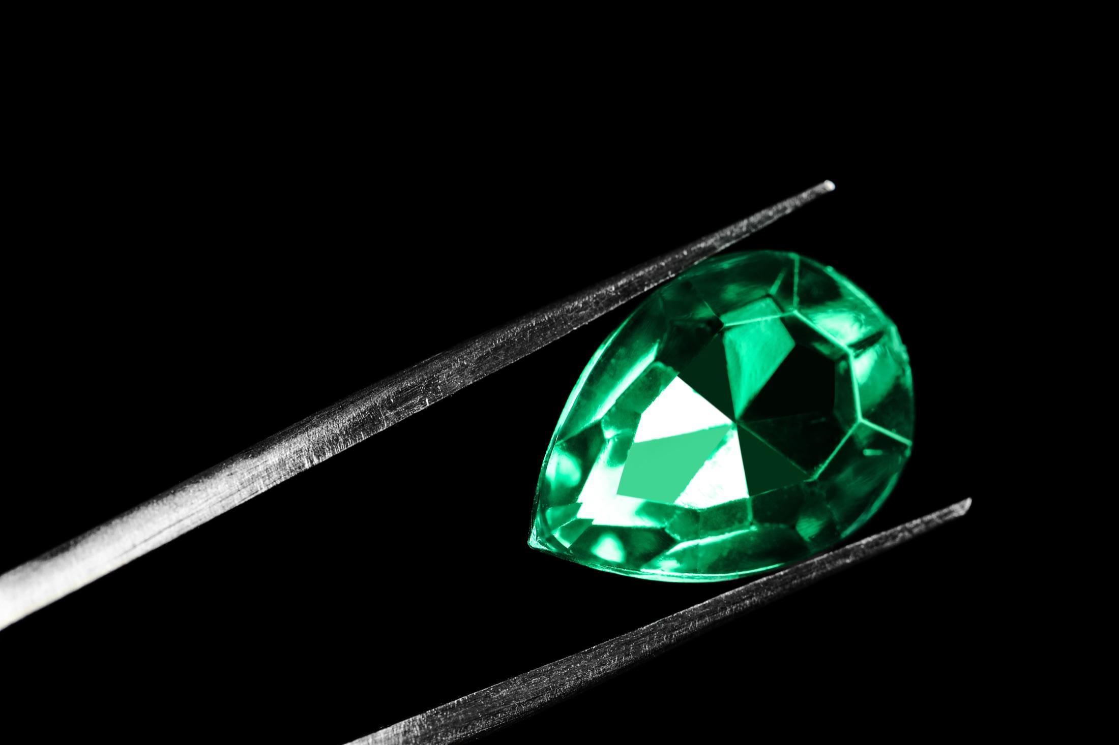 A pear-shaped emerald held with tweezers against a black background, presented for review